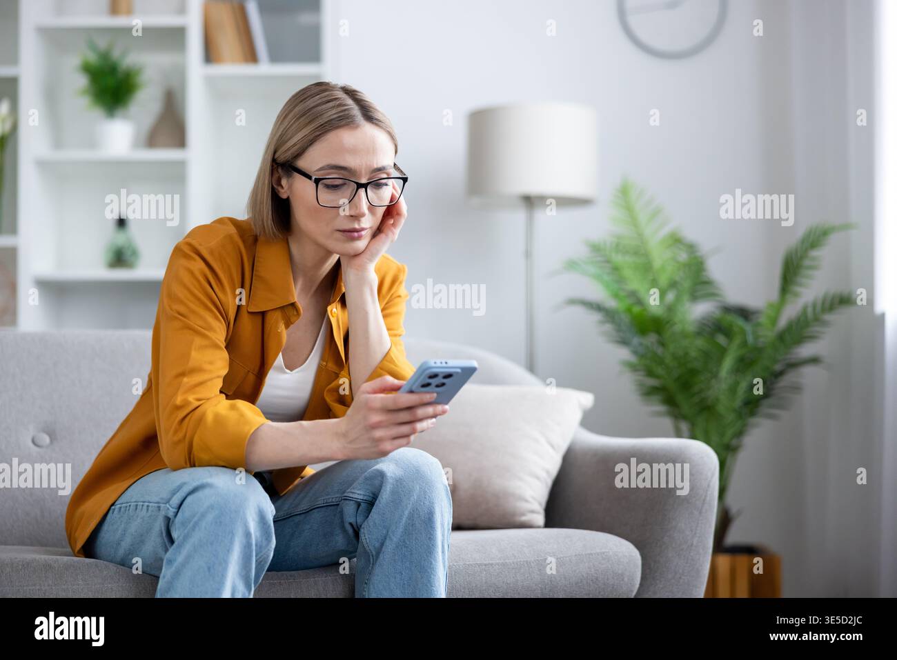 Femme dans des vêtements décontractés portant des lunettes assis sur un canapé gris tenant un smartphone, regardant l'écran avec une expression ennuyée et réfléchie, éprouvant la fatigue numérique dans un intérieur de la maison Banque D'Images