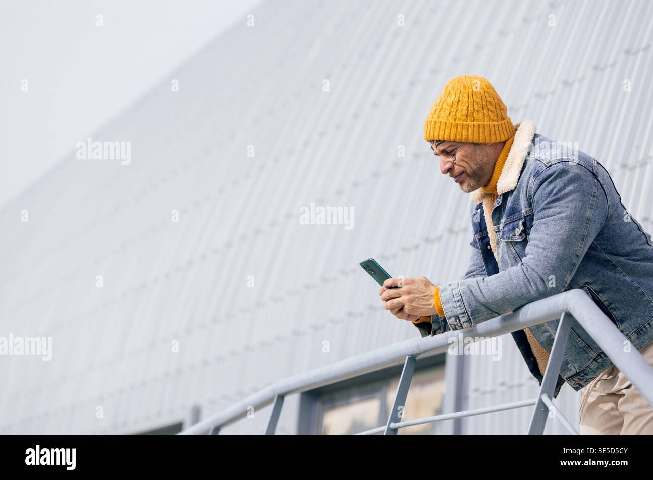 Homme décontracté avec un tatouage visage se penche sur une balustrade, vérifiant son téléphone tout en portant une veste en denim et un bonnet en tricot jaune dans un cadre urbain moderne. Banque D'Images