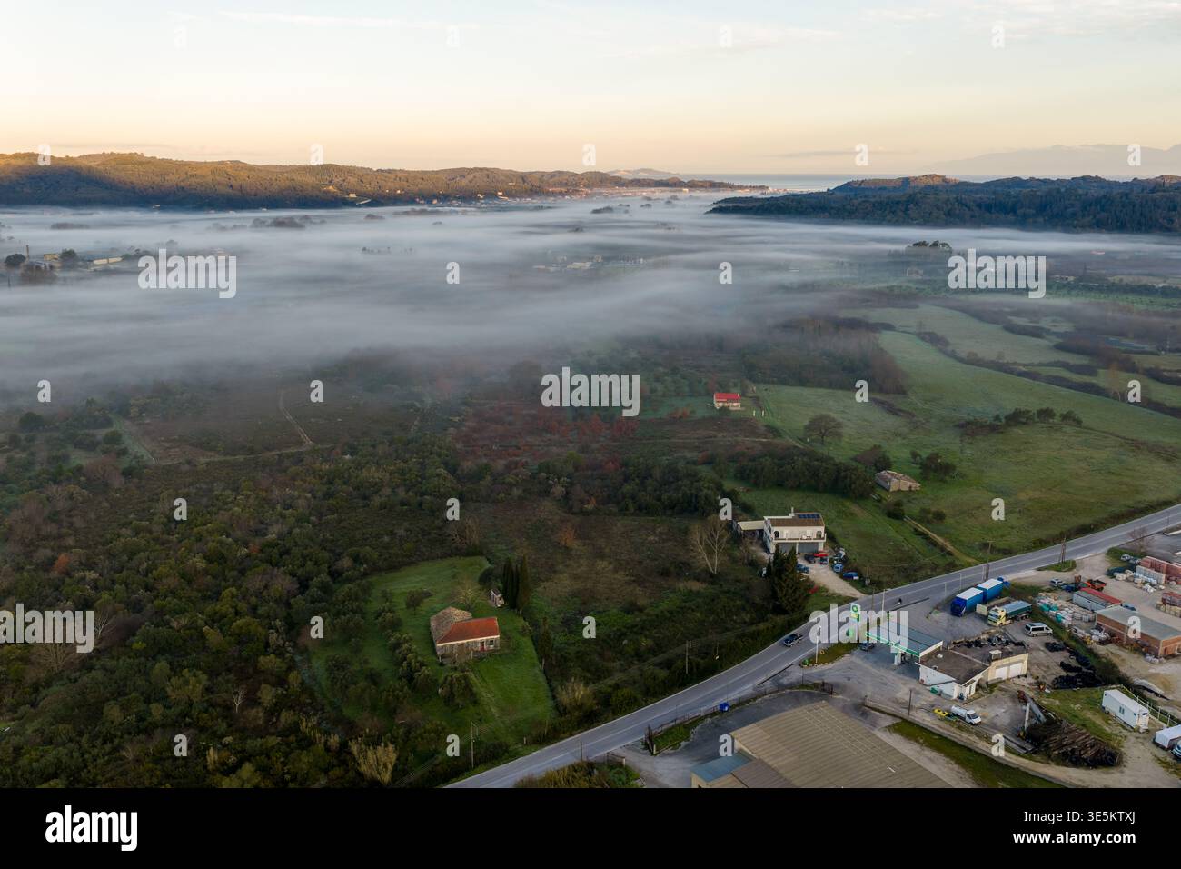 Paysage matinal aérien avec un brouillard épais couvrant une vallée, une route et des bâtiments ruraux pendant le lever du soleil dans le nord de Corfou, en Grèce Banque D'Images