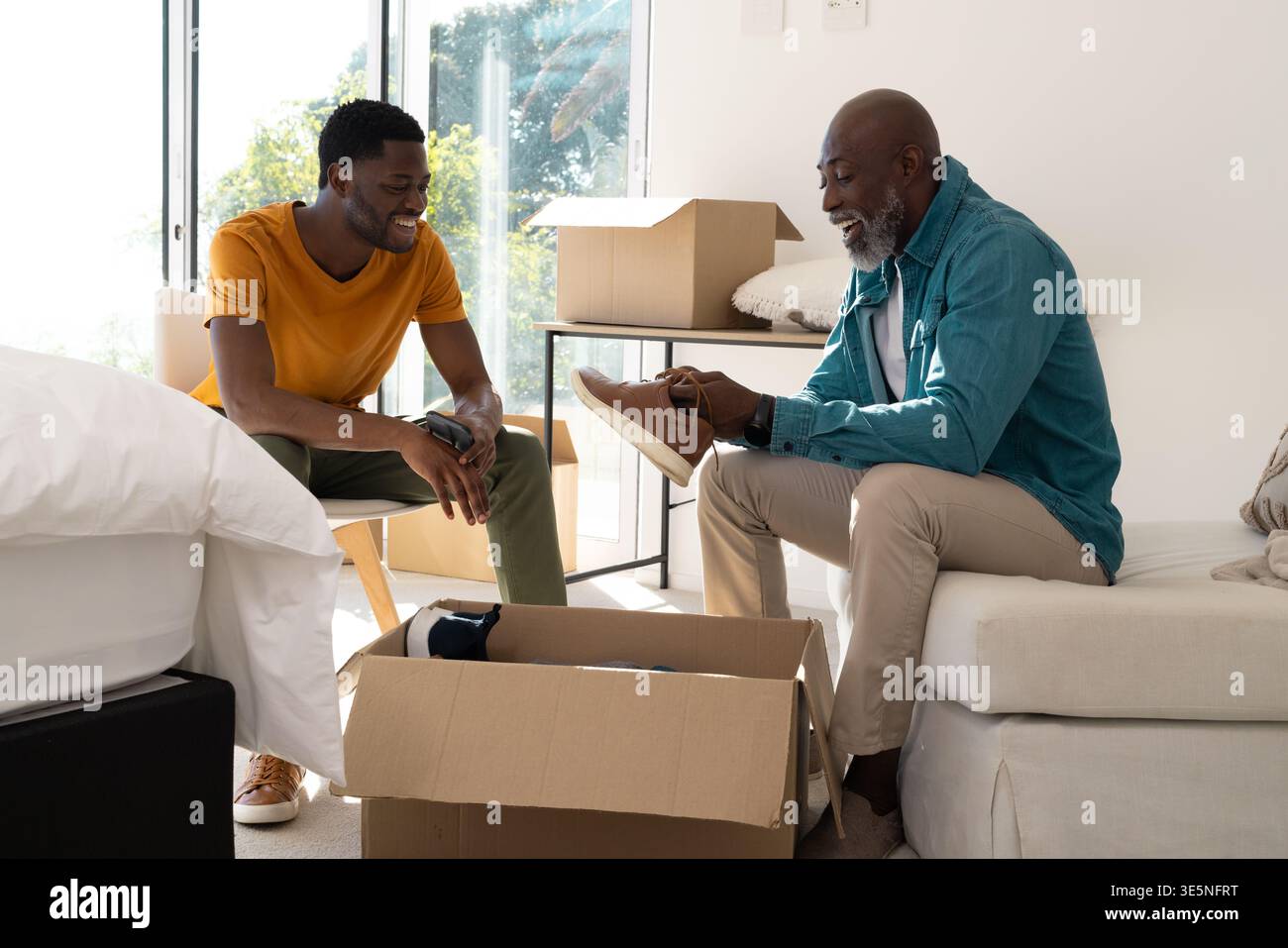 Déballer père et fils afro-américains à côté de la boîte de déménagement à la maison, tenant sneaker et smartphone Banque D'Images