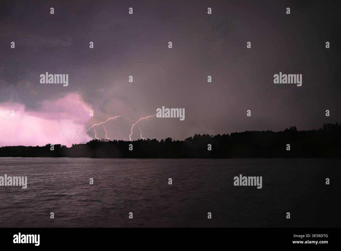 Plusieurs éclairs frappent au-delà d’une forêt sombre, éclairant les nuages d’orage et se reflétant faiblement sur la surface du lac la nuit. Banque D'Images