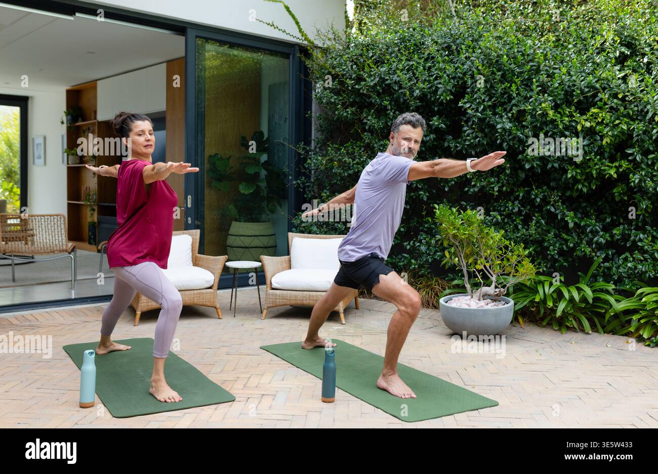 Couple diversifié pratiquant le yoga debout dans des vêtements de gymnastique sur le patio avec des tapis verts et des bouteilles d'eau Banque D'Images