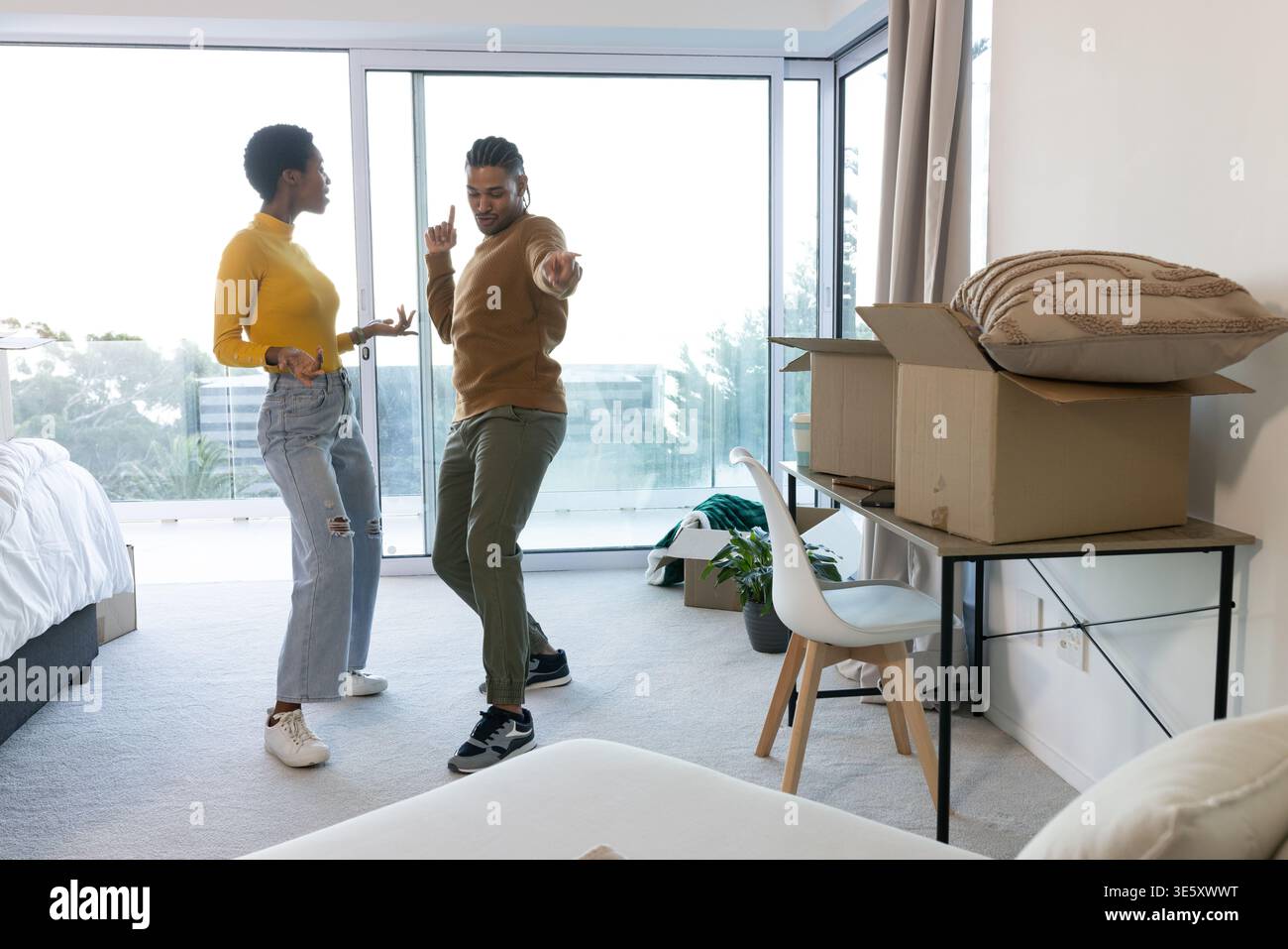 Danse couple diversifié filant dans la chambre avec portes de balcon, avec lit, boîtes de bureau et plante Banque D'Images