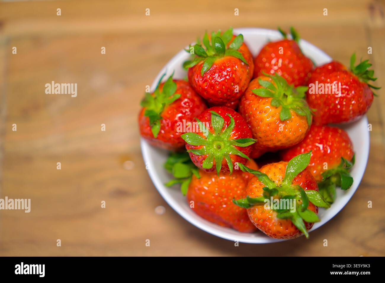 Vue en grand angle d'un bol en céramique blanche rempli à ras bord de grandes fraises mûres aux tiges vertes. Le bol repose sur un comptoir en bois naturel. La lumière du jour douce souligne la couleur rouge éclatante des baies et leur texture naturelle. fraises fraîches dans un bol blanc sur une surface en bois, concept alimentaire biologique. Photo de haute qualité Banque D'Images