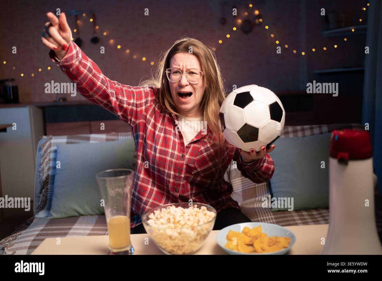 Émotionnellement fan de football féminin tenant le ballon regardant le match de football à la télévision à la maison assis sur le canapé avec la bière et des collations criant but. Sport, loisirs Banque D'Images