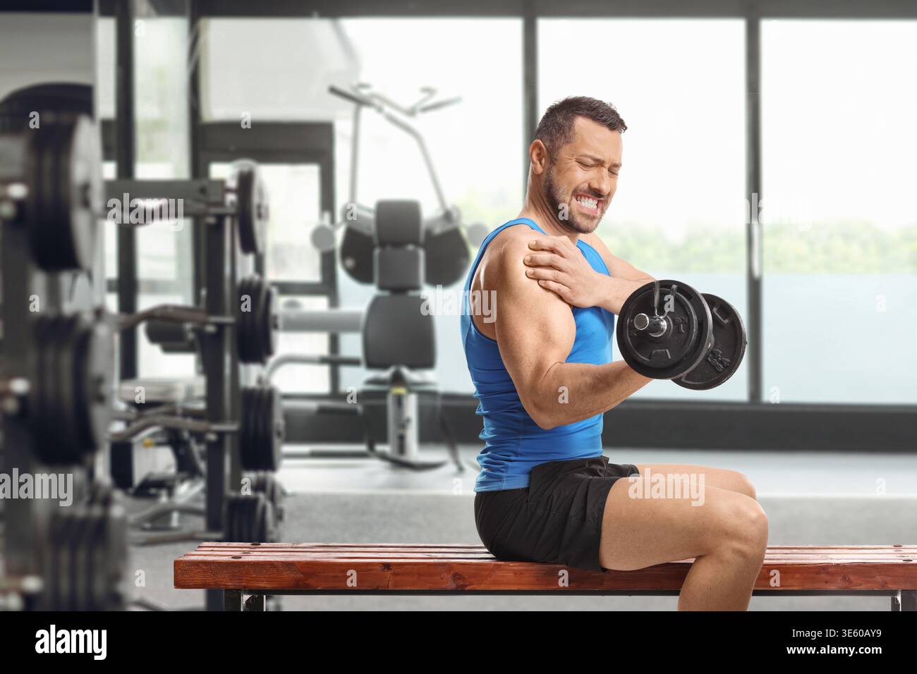 Homme dans un débardeur bleu ressentant la douleur à l'épaule tout en soulevant un haltère dans une salle de gym Banque D'Images