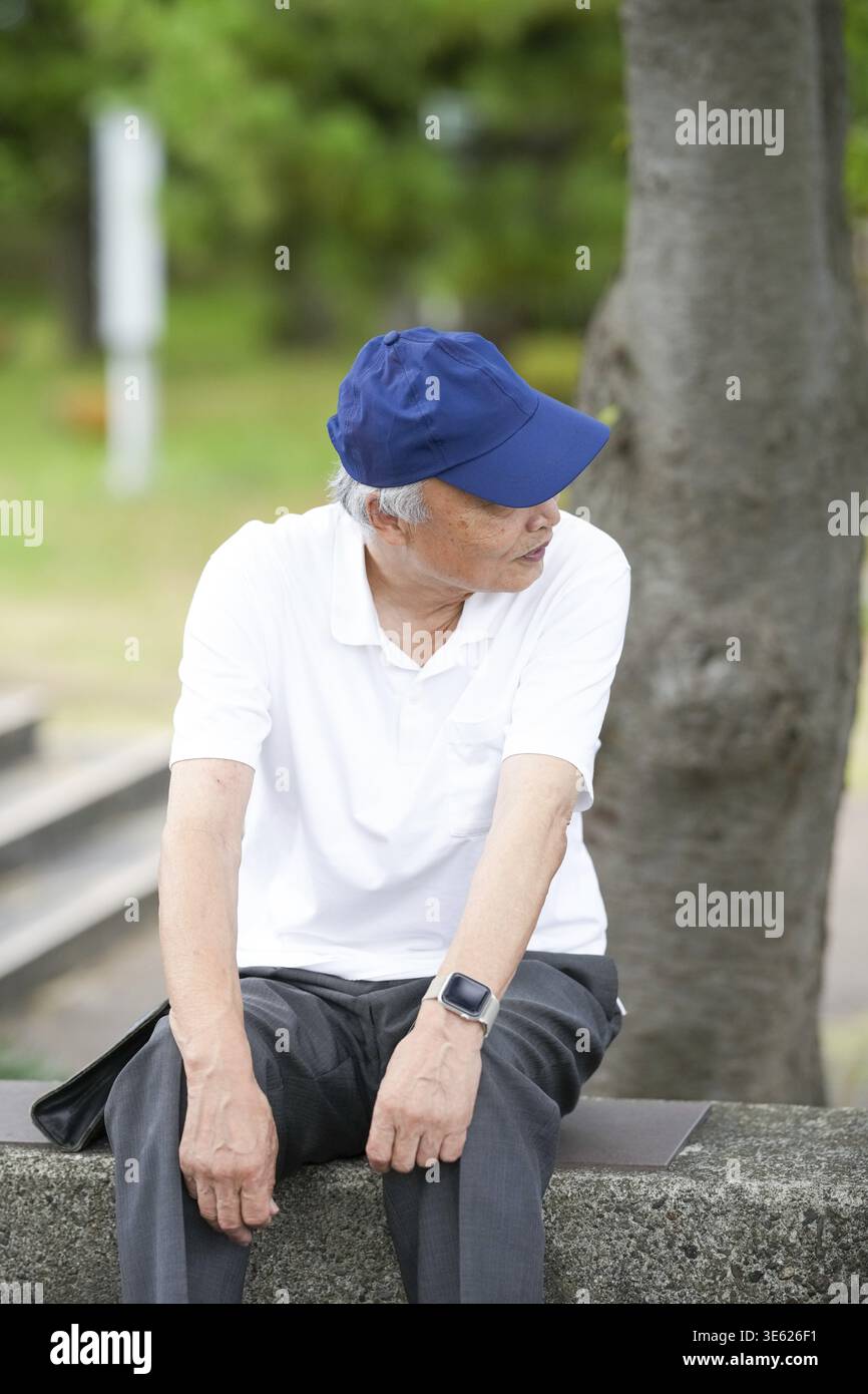 Un homme plus âgé portant un chapeau bleu et une chemise blanche est assis sur un banc. Il porte une montre et a une barbe grise Banque D'Images