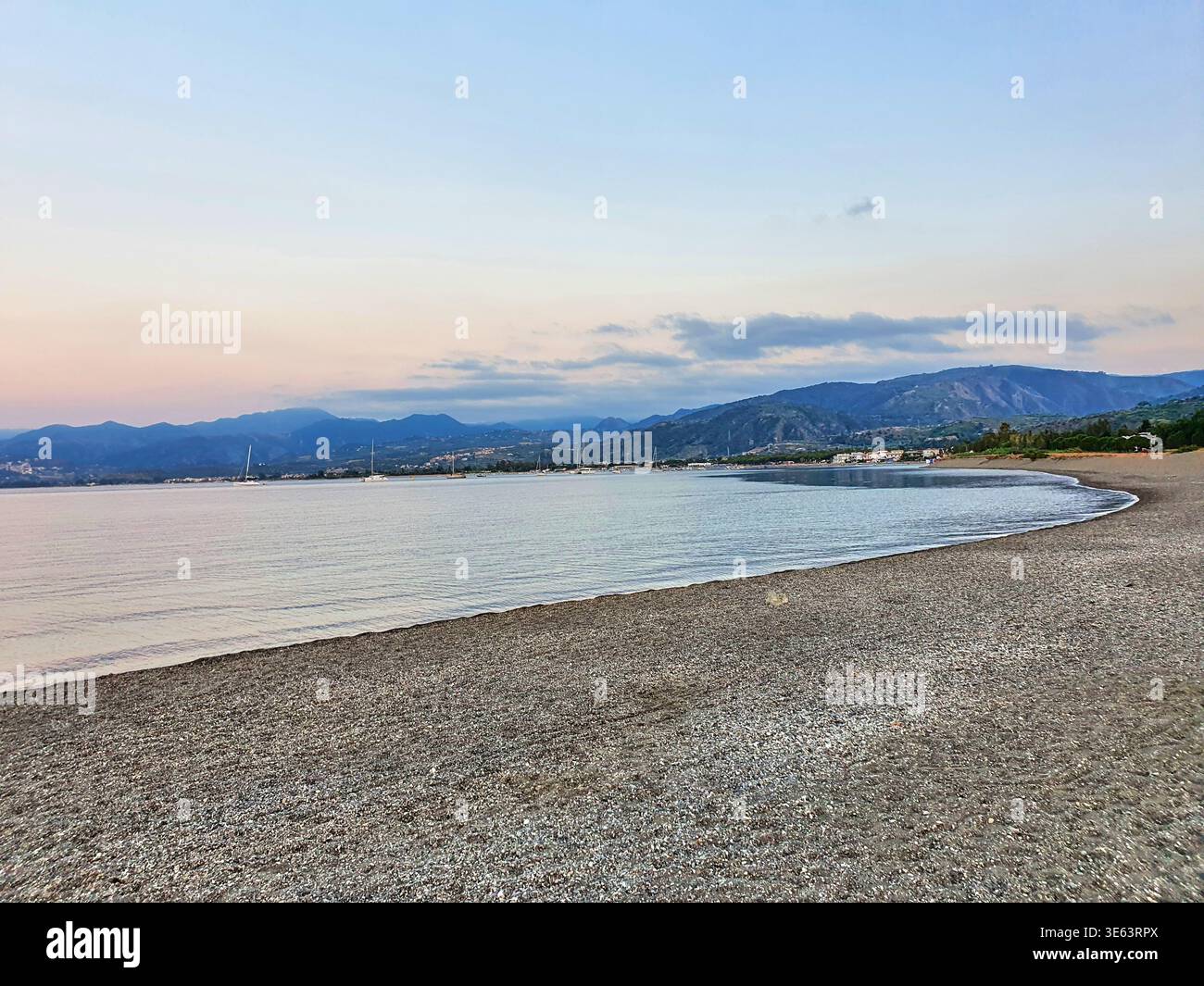 Un paysage côtier serein au crépuscule avec une plage de galets incurvée au premier plan avec de douces vagues lançant sur le rivage. La mer calme Banque D'Images