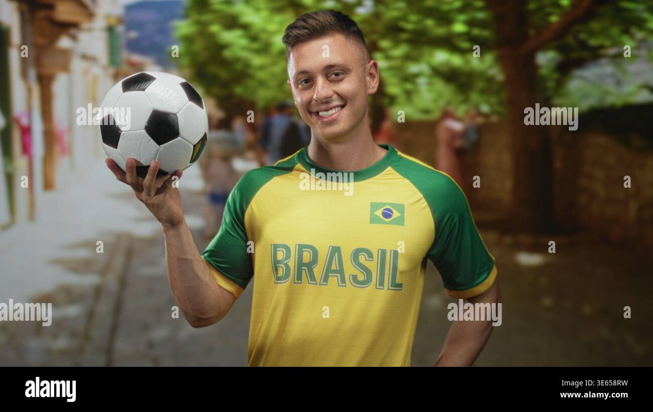 Man in brasil maillot tient le ballon de football avec la main droite et pointe le doigt tout en souriant dans la rue ; fierté joyeuse. Banque D'Images