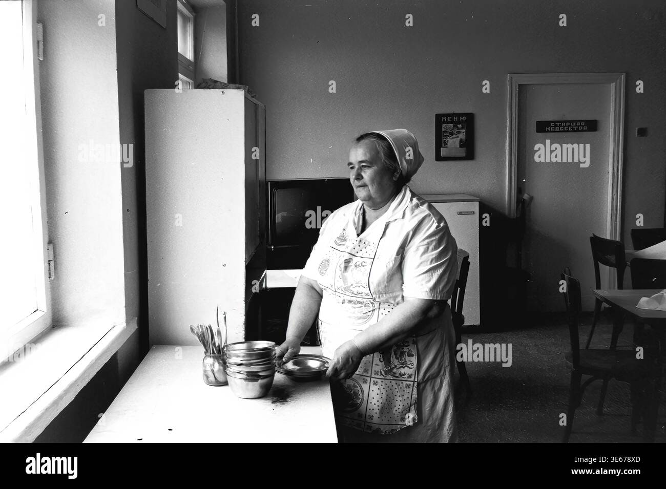 Sloviansk, 1983 ans, hôpital AIZ. Une employée d'hôpital dans un uniforme blanc et un tablier à motifs se tient près d'une table dans la salle à manger, tenant soigneusement et empilant des bols en métal. La chambre présente des détails intérieurs typiquement soviétiques, y compris un téléviseur, un réfrigérateur et un panneau de porte indiquant la lecture de l'infirmière en chef. Cette image d'archives authentique met en évidence la routine quotidienne, les services de restauration et l'infrastructure domestique au sein d'un établissement médical, préservant un moment authentique de travail et de vie quotidienne dans le Donbass paisible. Banque D'Images