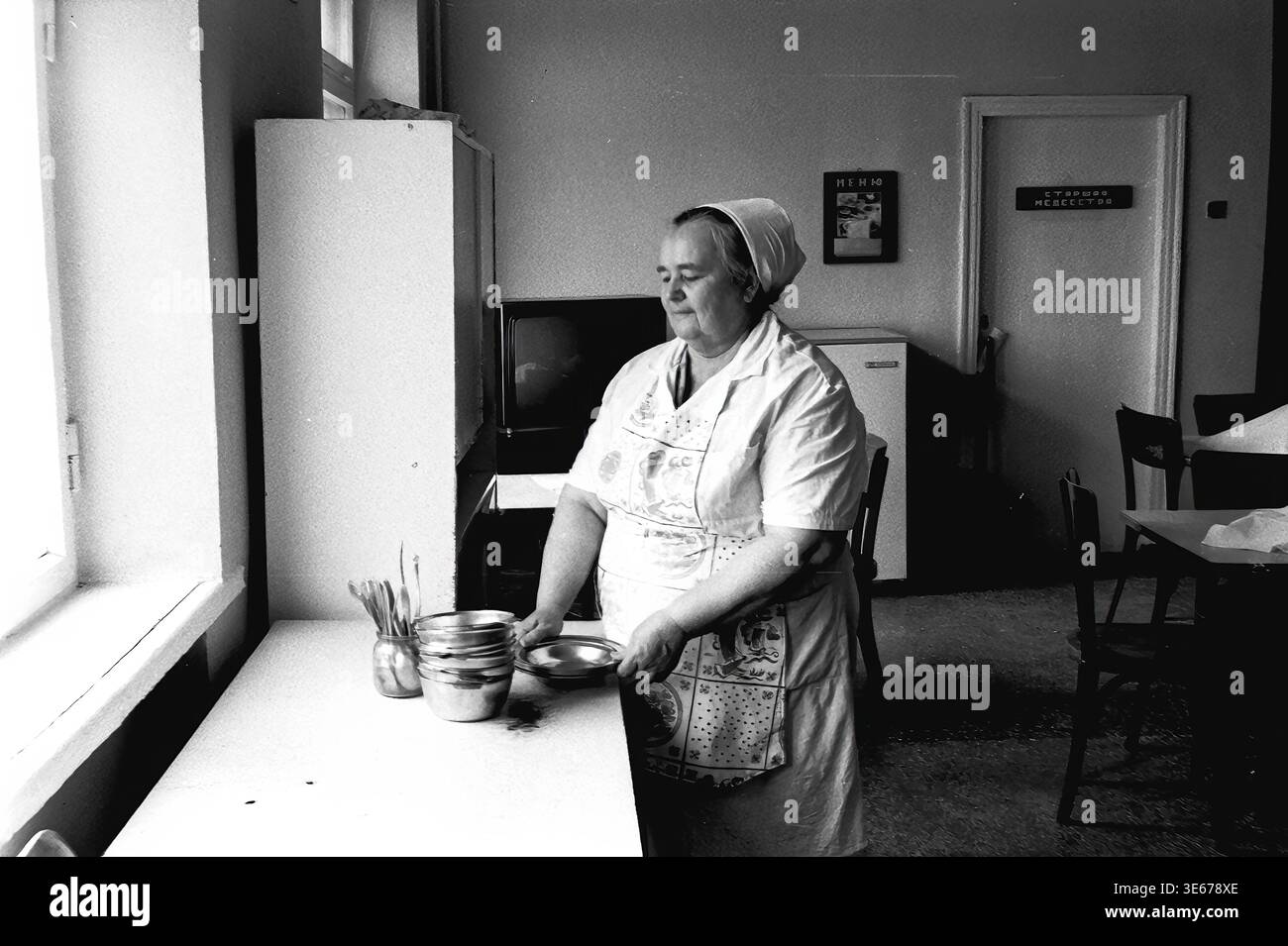 Sloviansk, 1983 ans, hôpital AIZ. Une employée d'hôpital dans un uniforme blanc et un tablier à motifs se tient près d'une table dans la salle à manger, tenant soigneusement et empilant des bols en métal. La chambre présente des détails intérieurs typiquement soviétiques, y compris un téléviseur, un réfrigérateur et un panneau de porte indiquant la lecture de l'infirmière en chef. Cette image d'archives authentique met en évidence la routine quotidienne, les services de restauration et l'infrastructure domestique au sein d'un établissement médical, préservant un moment authentique de travail et de vie quotidienne dans le Donbass paisible. Banque D'Images