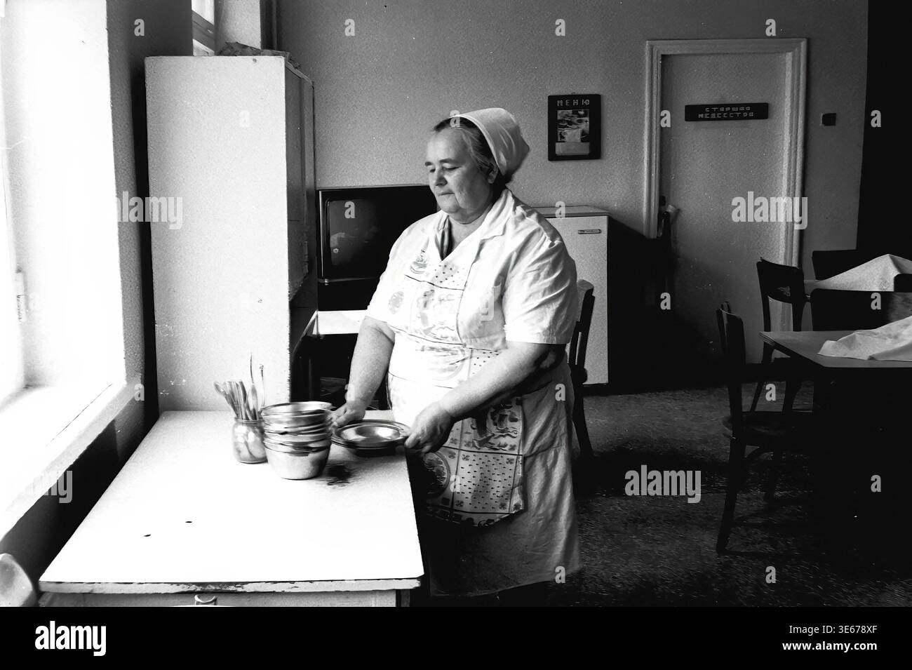 Sloviansk, 1983 ans, hôpital AIZ. Une employée d'hôpital dans un uniforme blanc et un tablier à motifs se tient près d'une table dans la salle à manger, tenant soigneusement et empilant des bols en métal. La chambre présente des détails intérieurs typiquement soviétiques, y compris un téléviseur, un réfrigérateur et un panneau de porte indiquant la lecture de l'infirmière en chef. Cette image d'archives authentique met en évidence la routine quotidienne, les services de restauration et l'infrastructure domestique au sein d'un établissement médical, préservant un moment authentique de travail et de vie quotidienne dans le Donbass paisible. Banque D'Images