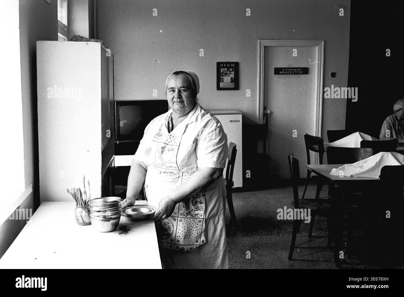 Sloviansk, 1983 ans, hôpital AIZ. Une employée d'hôpital dans un uniforme blanc et un tablier à motifs se tient près d'une table dans la salle à manger, tenant soigneusement et empilant des bols en métal. La chambre présente des détails intérieurs typiquement soviétiques, y compris un téléviseur, un réfrigérateur et un panneau de porte indiquant la lecture de l'infirmière en chef. Cette image d'archives authentique met en évidence la routine quotidienne, les services de restauration et l'infrastructure domestique au sein d'un établissement médical, préservant un moment authentique de travail et de vie quotidienne dans le Donbass paisible. Banque D'Images
