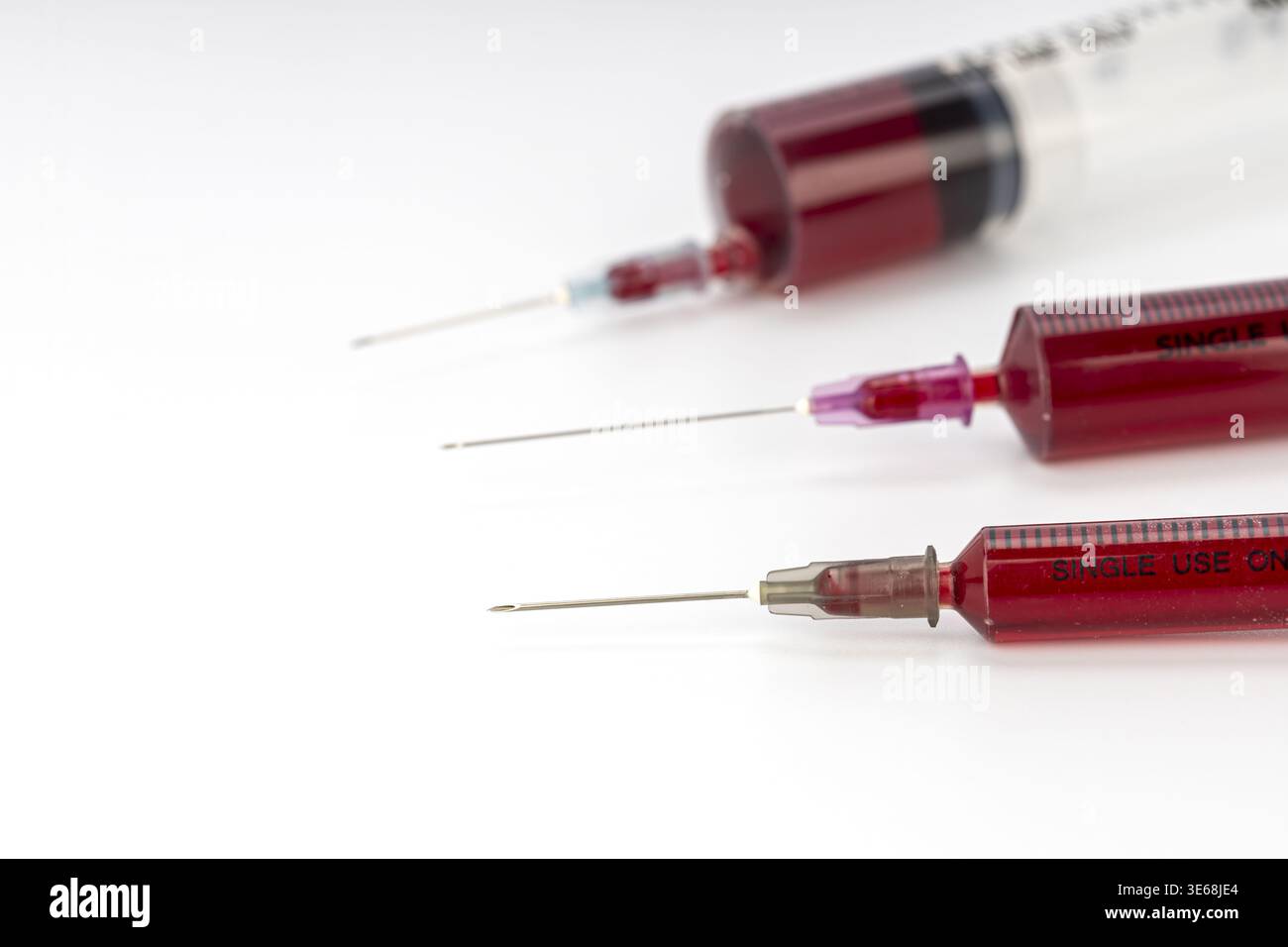 Trois seringues médicales à usage unique remplies de sang rouge foncé reposent sur un fond blanc propre. Les aiguilles exposées et les barils en plastique étiquetés répètent Banque D'Images