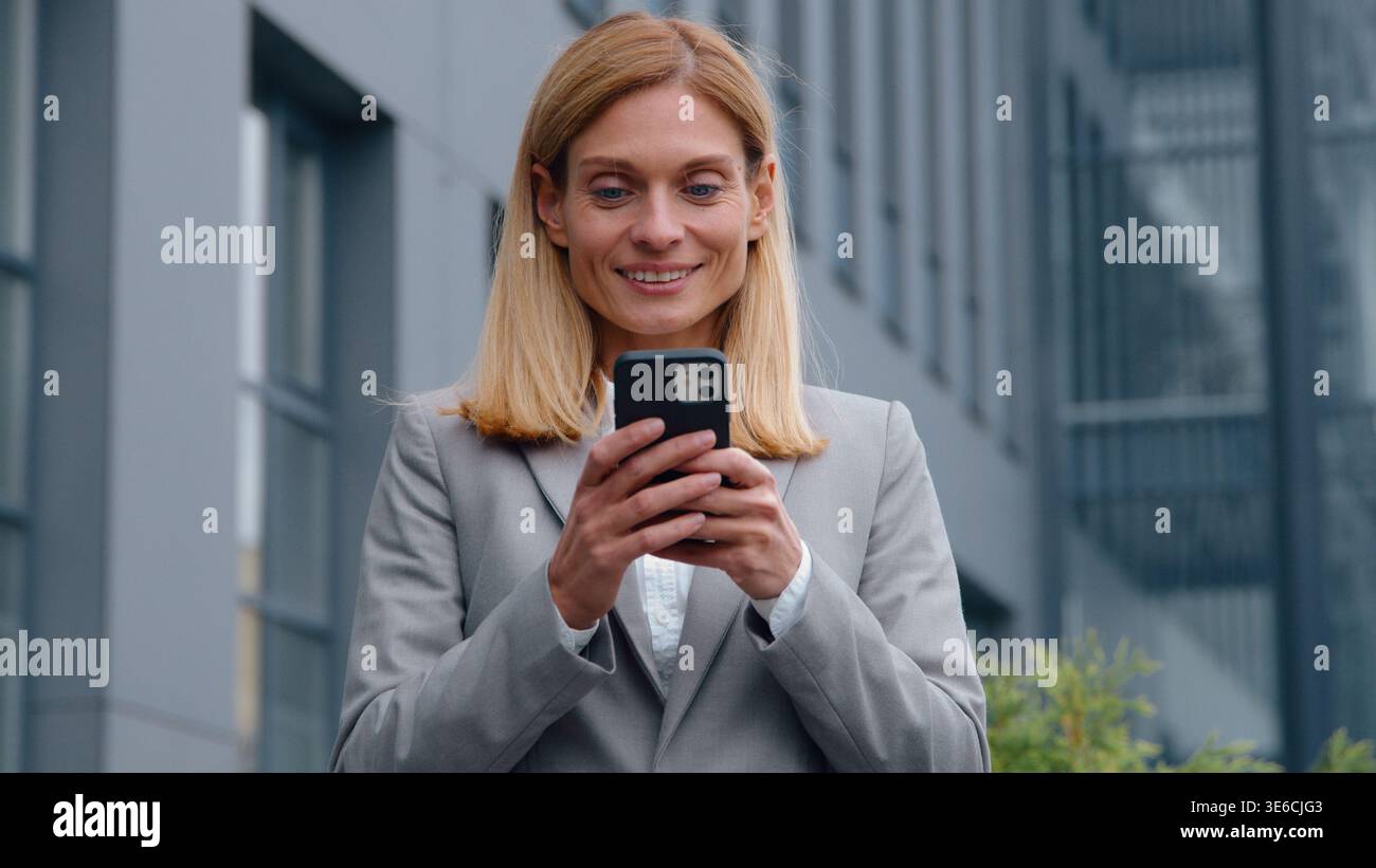 Heureuse femme d'affaires adulte caucasienne avec le téléphone appréciant le contenu en ligne dans la ville femme d'affaires d'âge moyen discutant le message électronique exécutif féminin Banque D'Images