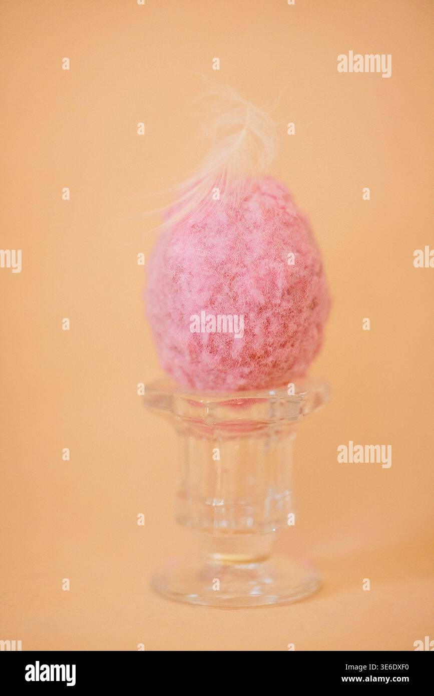 Oeuf de Pâques en feutre rose avec une plume blanche affichée sur un piédestal en verre, sur un fond orange doux pour un décor de vacances de printemps Banque D'Images