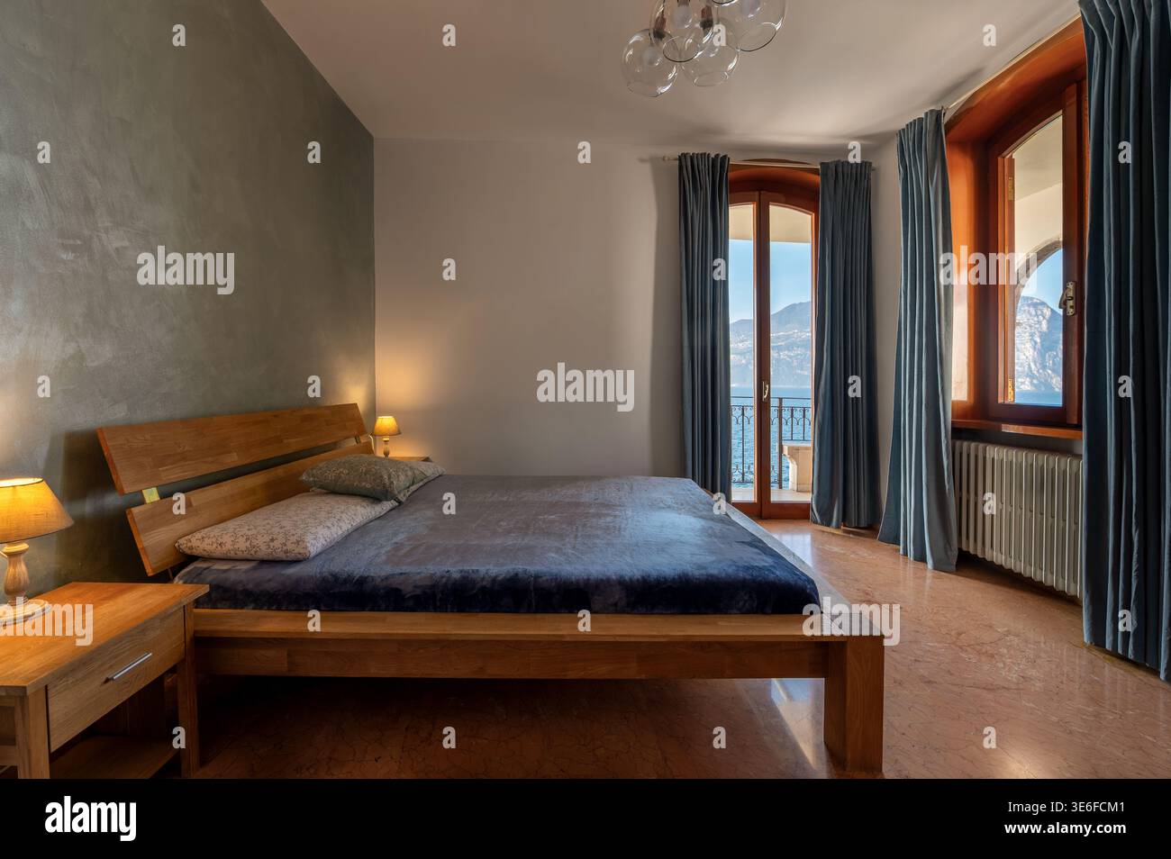 Élégante villa de luxe chambre avec lit en bois, éclairage tamisé et de grandes fenêtres ouvrant sur un balcon avec vue sereine sur le lac et la montagne, idéal pour un Banque D'Images