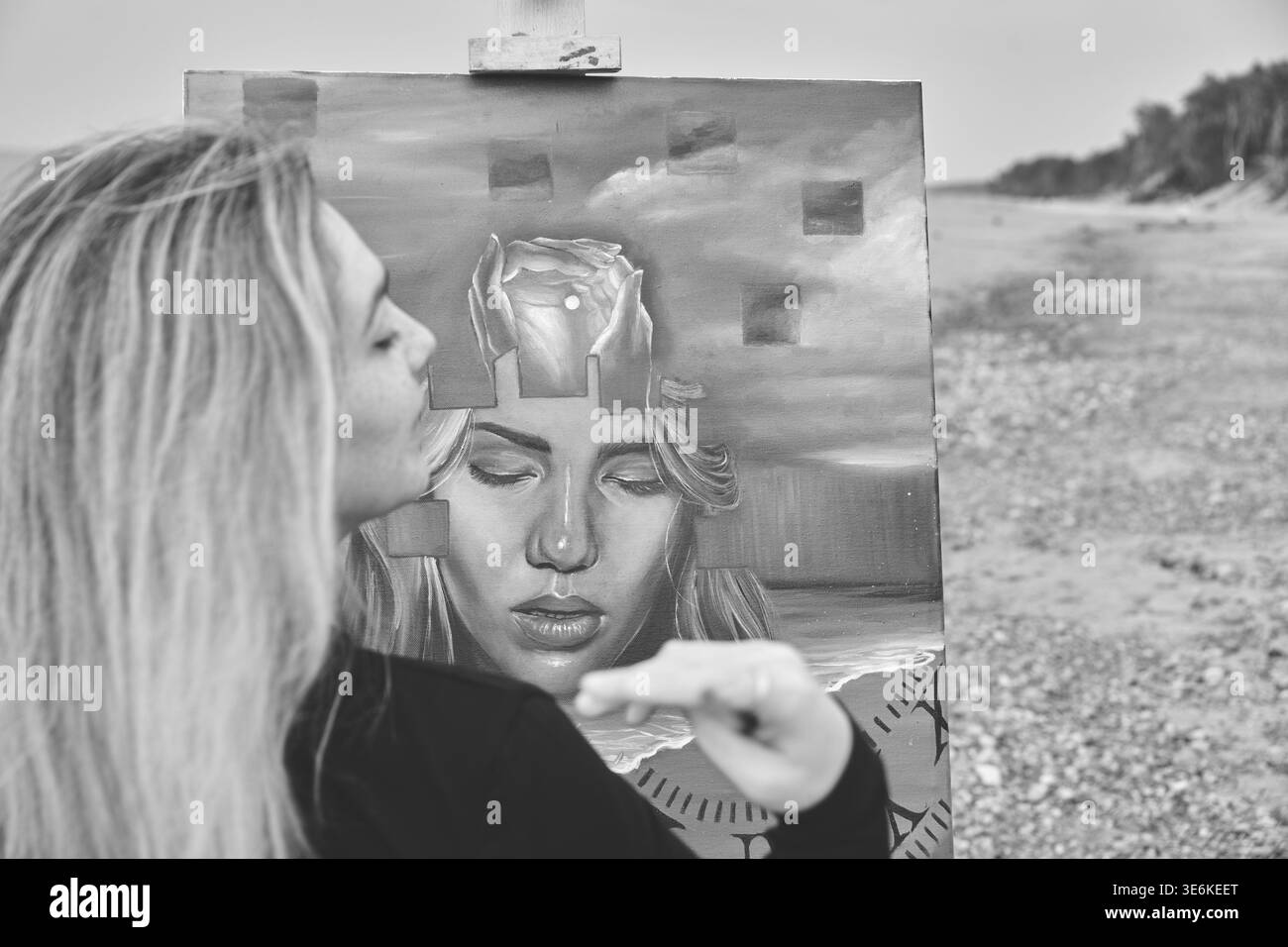 Artiste femme adulte avec de longs cheveux blonds créant autoportrait surréaliste sur toile, situé sur la plage de galets avec la mer calme et les arbres en arrière-plan und Banque D'Images