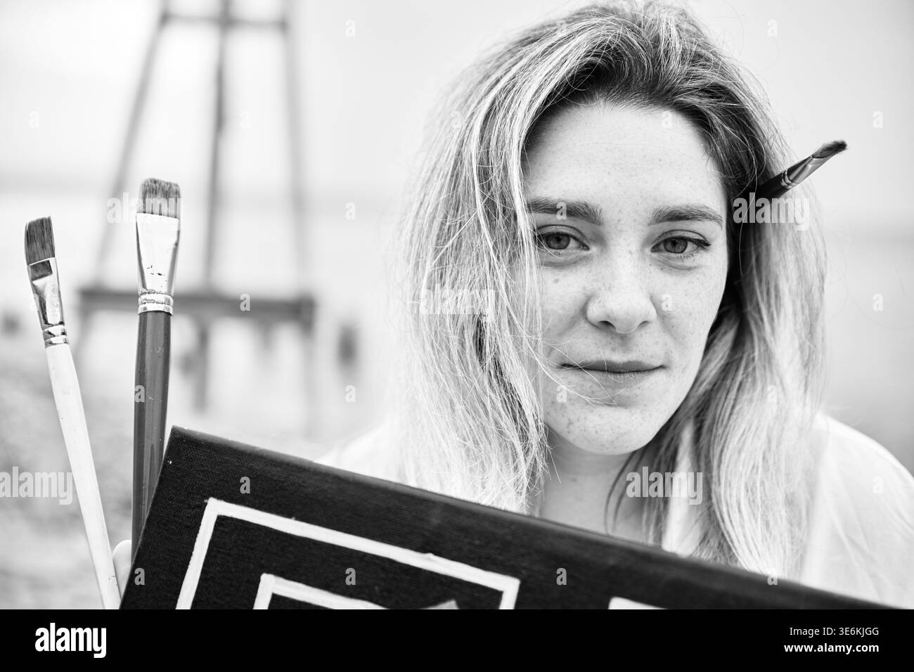 Femme adulte artiste avec les cheveux clairs et le sourire lent tient la peinture et les pinceaux, capturé gros plan dans un éclairage naturel doux avec arrière-plan de studio flou Banque D'Images