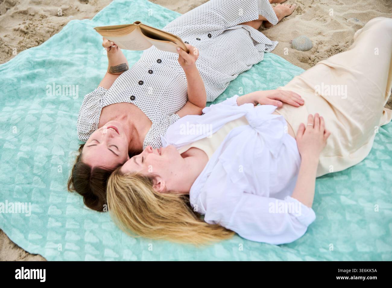 Deux femmes adultes allongées sur une couverture bleu clair sur Sandy Beach lisant paisiblement le livre ensemble sous une lumière naturelle douce. La scène transmet le calme, le loisir, et Banque D'Images
