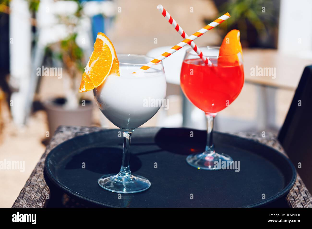 Deux cocktails colorés sont assis sur un plateau noir, un blanc et un rouge, chacun garni d'une tranche d'orange et de paille rayée. La scène lumineuse évoque un re Banque D'Images