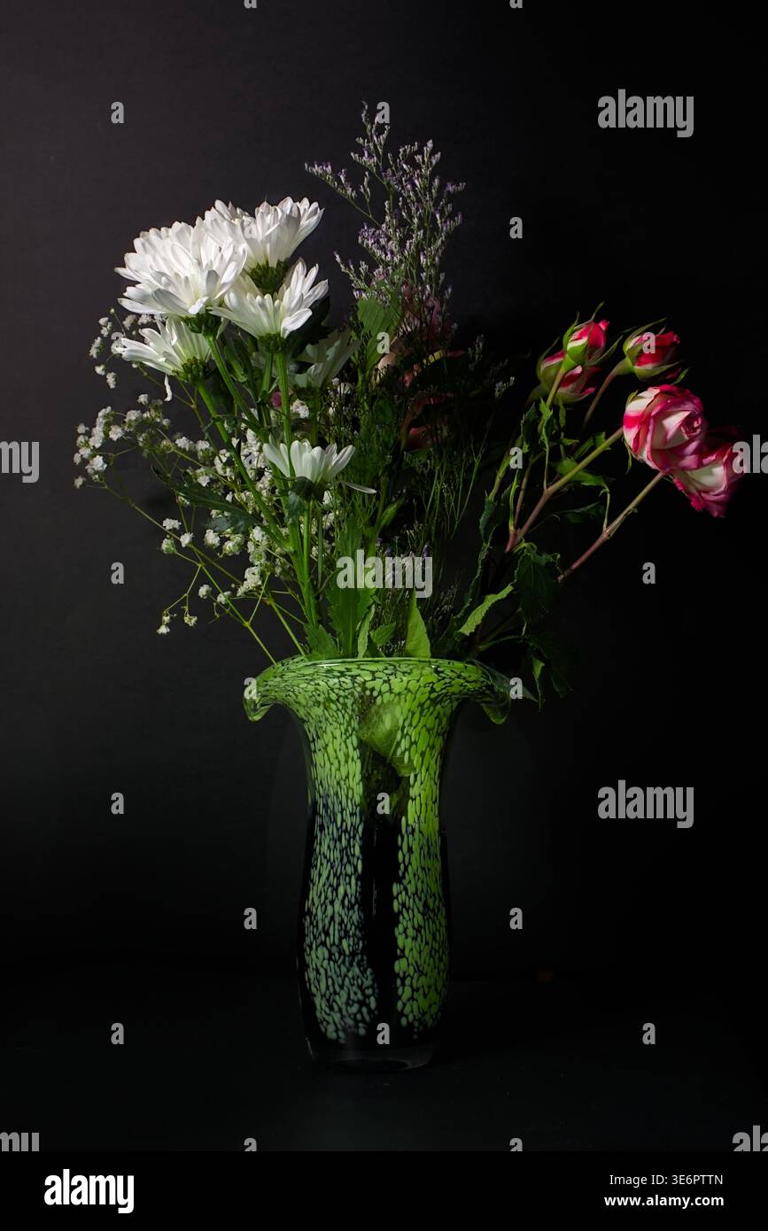 Arrangement floral élégant avec marguerites blanches et roses rouges dans un vase en verre d'art tacheté vert sur noir Banque D'Images