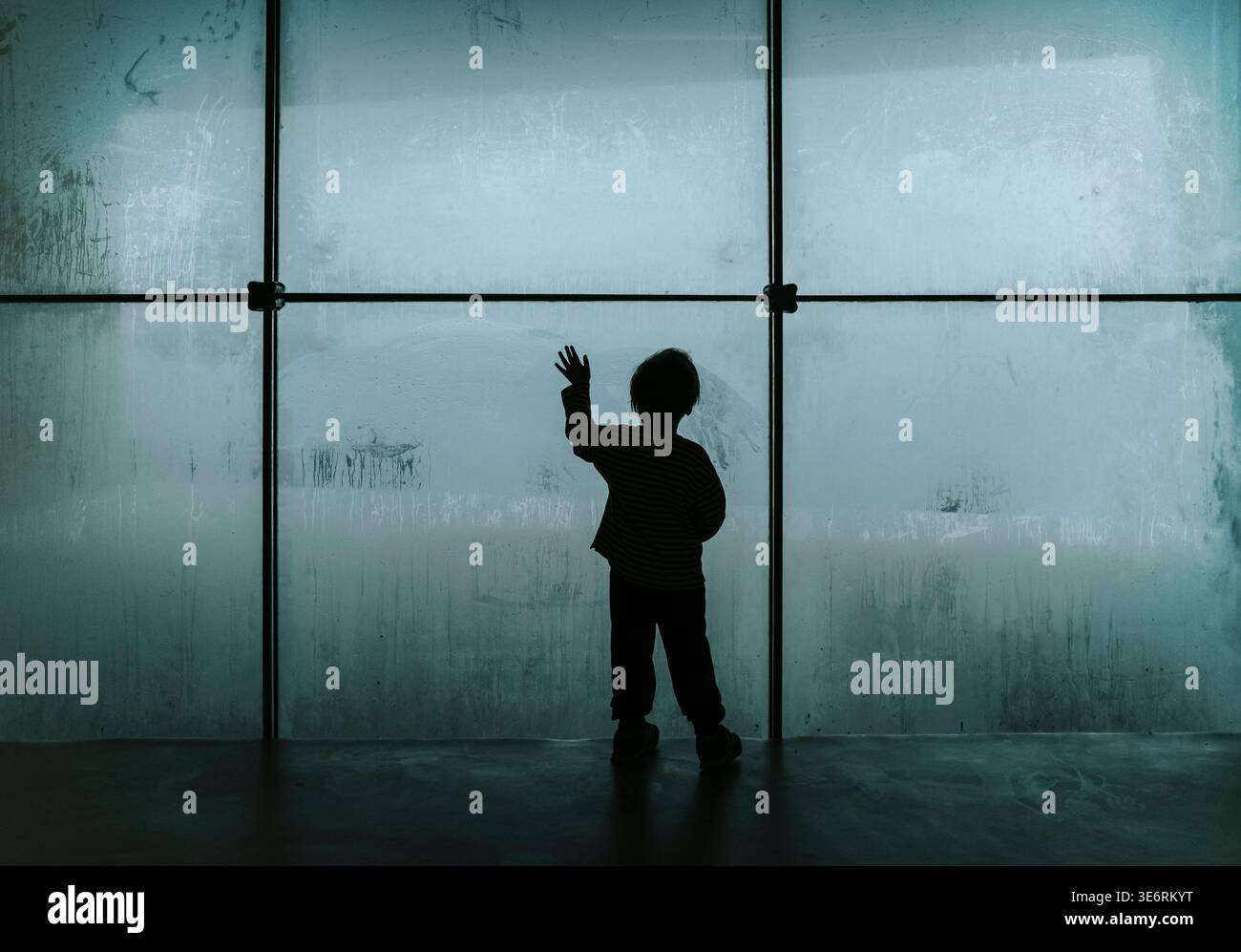 Silhouette d'un enfant solitaire regardant à travers une grande fenêtre embuée froide. Un travail de photographie conceptuelle explorant les thèmes de la dépression infantile, de la crise psychologique et du profond sentiment d’isolement et de tristesse. Photo de haute qualité Banque D'Images