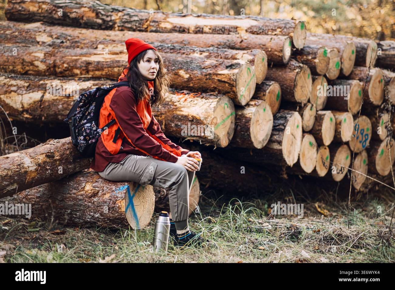 Femme avec sac à dos est assis sur la bûche tombée tenant la tasse près de la pile de bois. détox numérique nature, santé mentale en plein air, évasion en pleine nature. Banque D'Images