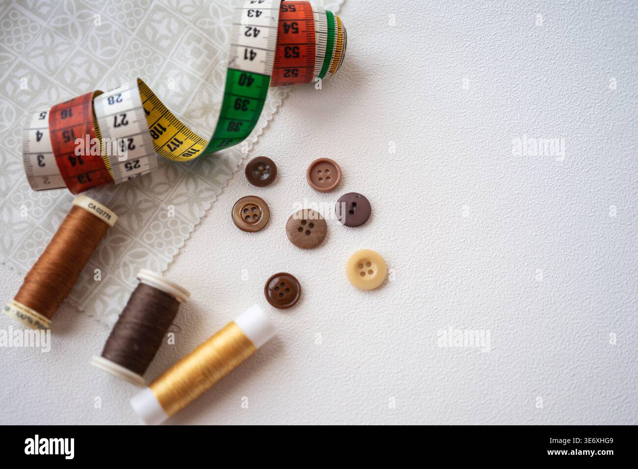 Une composition de couture avec un ruban à mesurer enroulé, des bobines de fil en sha-des de brun et d'or, et des boutons assortis sur un dos clair et texturé Banque D'Images