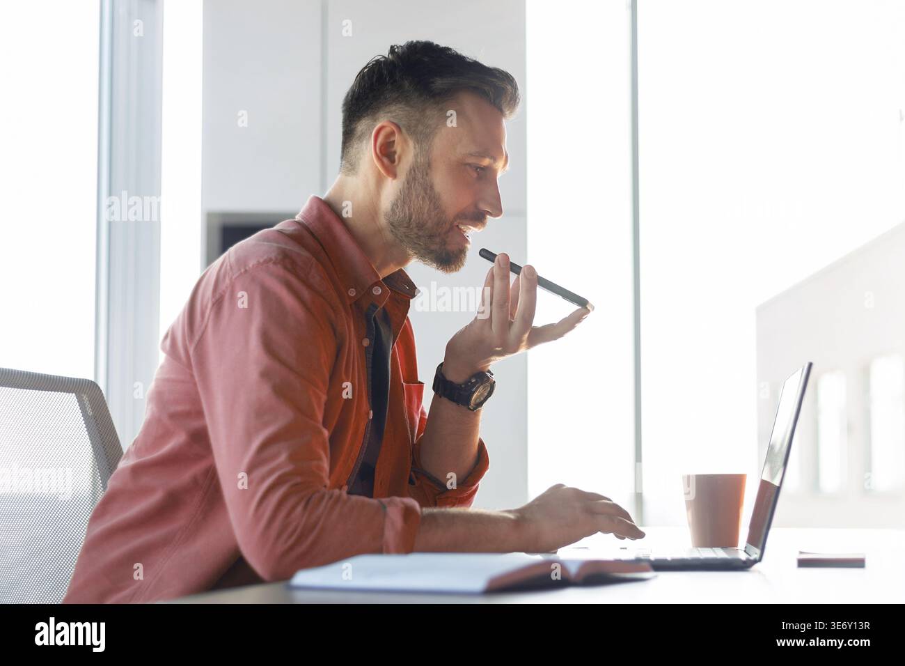 Homme utilisant la commande vocale sur ordinateur portable tout en étant assis à un bureau dans une pièce lumineuse Banque D'Images