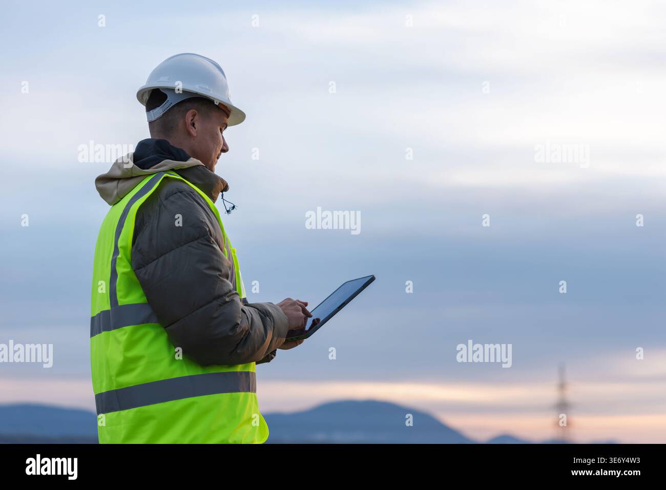 Un jeune ingénieur électricien travaille à l'extérieur, inspectant des lignes électriques à haute tension avec une tablette dans une zone de service public au crépuscule. Banque D'Images
