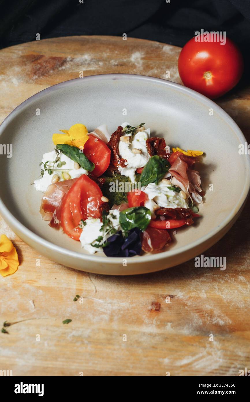 Salade Caprese italienne gastronomique avec fromage burrata frais, jambon prosciutto, tomates cerises et sauce pesto garnie de fleurs comestibles. Banque D'Images