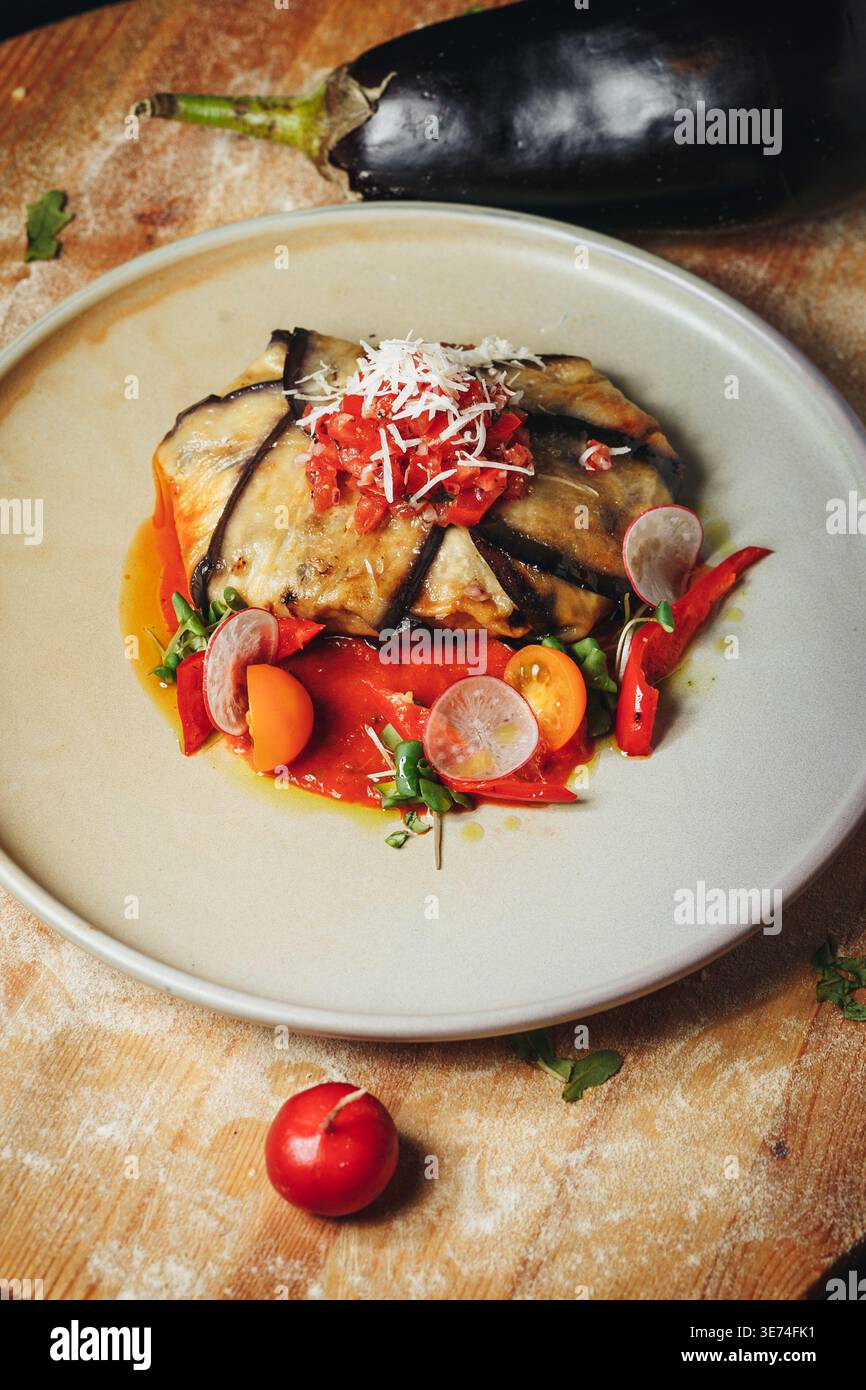 Roulés gourmands d'aubergines rôties servis sur une sauce au poivron rouge éclatante avec tomates cerises fraîches et garniture de radis sur une assiette en céramique. Banque D'Images