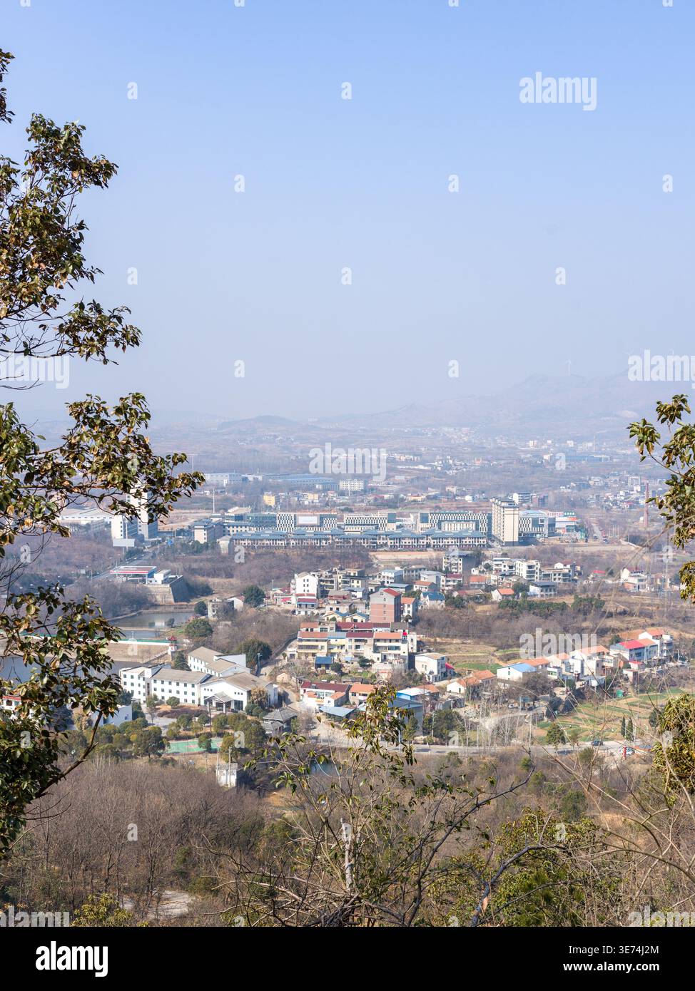 Une vue panoramique surélevée depuis une colline boisée surplombant une ville lointaine ou une ville située dans une vallée, encadrée par des arbres sous un ciel bleu brumeux. La scène R Banque D'Images
