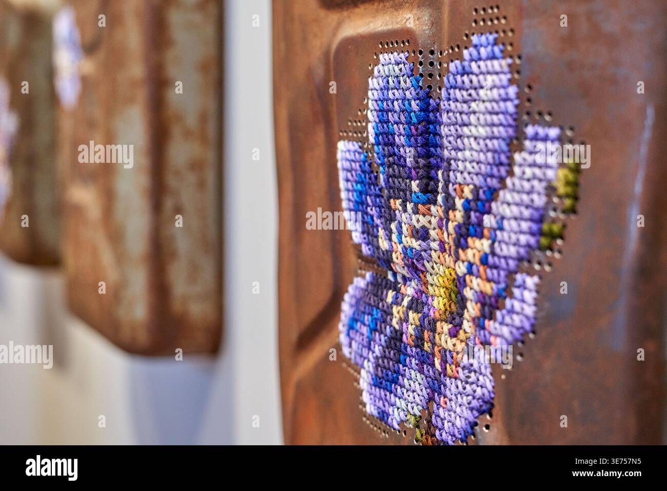 Photographie en gros plan d'une œuvre d'art textile contemporaine présentant une broderie au point de croix d'une fleur violette cousue sur une surface métallique rouillée. La Banque D'Images