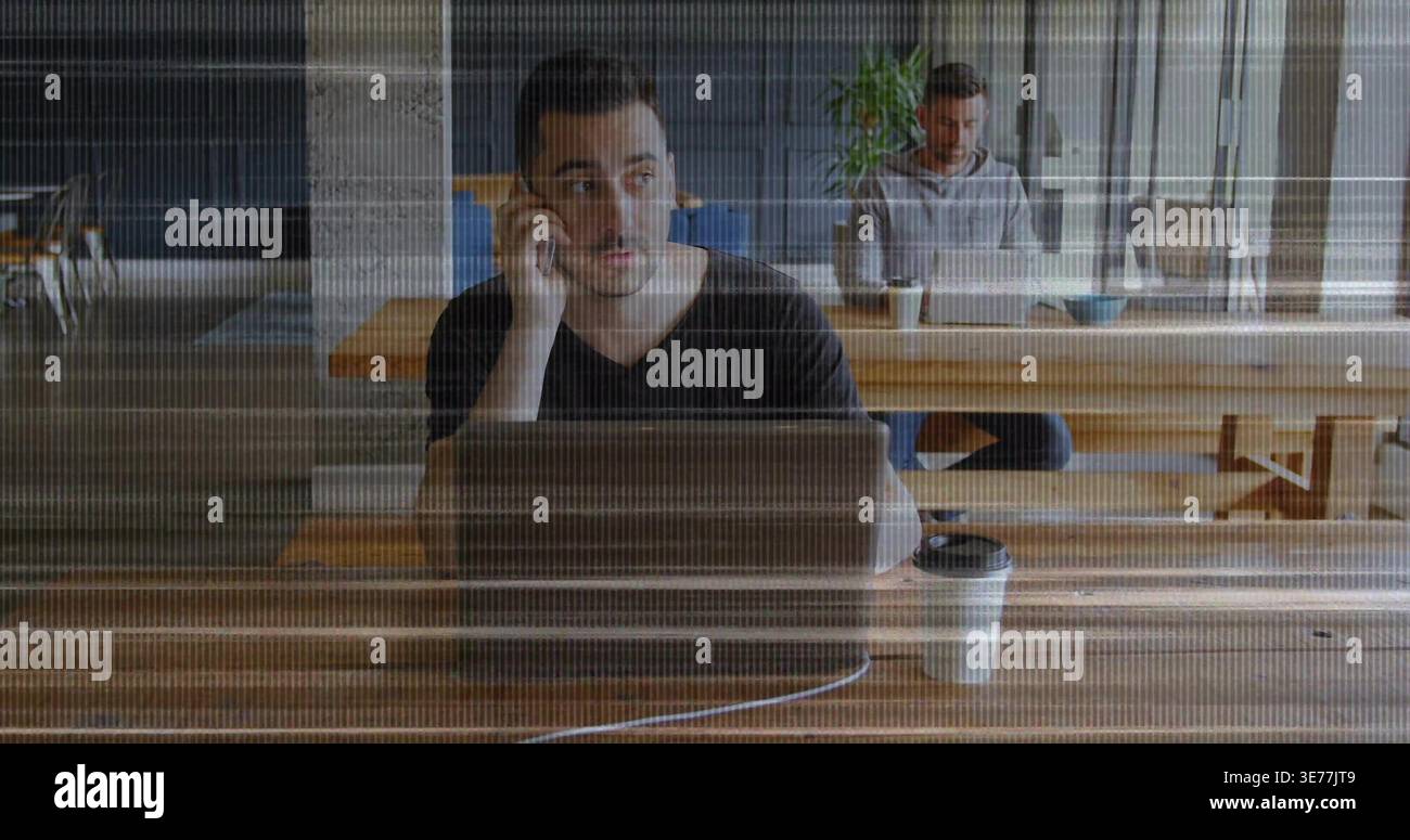Deux hommes mi-adultes partageant la table commune, l'homme de devant qui sonne le téléphone pour le travail sur ordinateur portable Banque D'Images