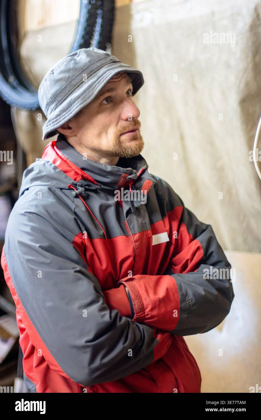 Un homme pensif d'âge moyen portant une veste d'hiver chaude et un chapeau seau se tient debout avec les bras croisés dans un cadre d'atelier, regardant hors caméra. Banque D'Images