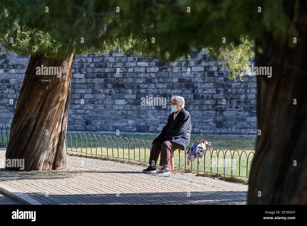 Homme âgé portant un masque médical assis sur un tabouret contre un mur de pierre dans un parc public, Pékin, Chine, Asie. Banque D'Images