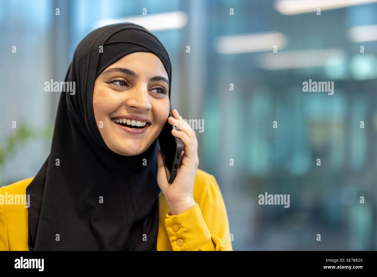 Femme musulmane en hijab et chemise jaune ayant une conversation joyeuse sur son téléphone portable, communiquant activement tout en travaillant dans un bureau moderne envir Banque D'Images