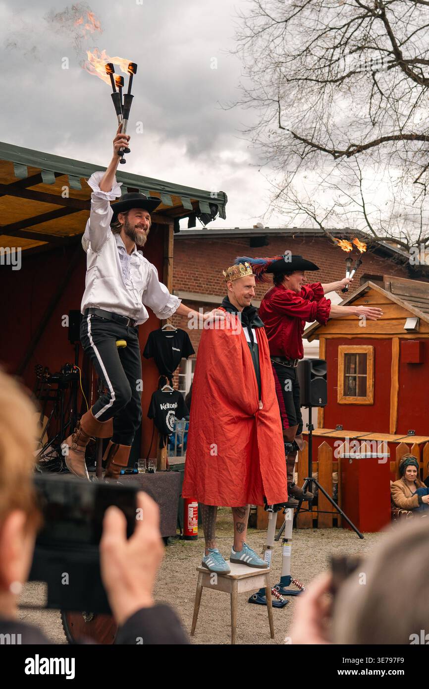 Berlin, Allemagne - 5 avril 2026 : des artistes de feu en costumes de théâtre se tiennent debout sur de grandes monocycles à côté d'un volontaire couronné lors d'un concert sur scène à Banque D'Images