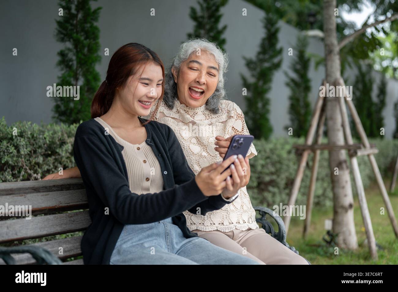 Femme thaïlandaise ayant du plaisir à partager smartphone avec sa grand-mère tout en étant assise sur un banc en bois dans le jardin pendant la journée Banque D'Images