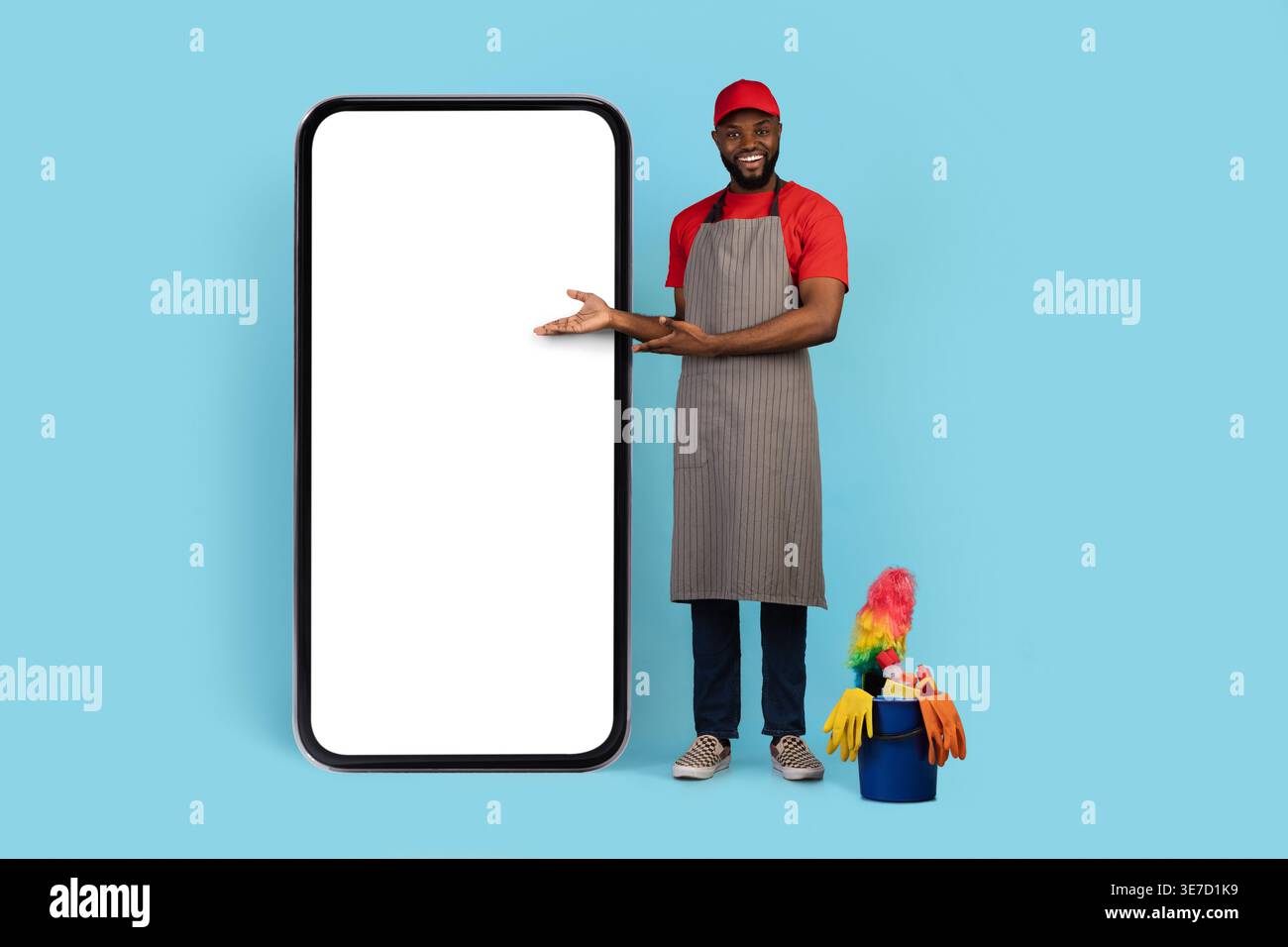 Capuchon de port de nettoyant masculin noir et tablier pointant les mains sur le grand smartphone debout près du panier d'outils de nettoyage sur fond bleu dans le studio Banque D'Images