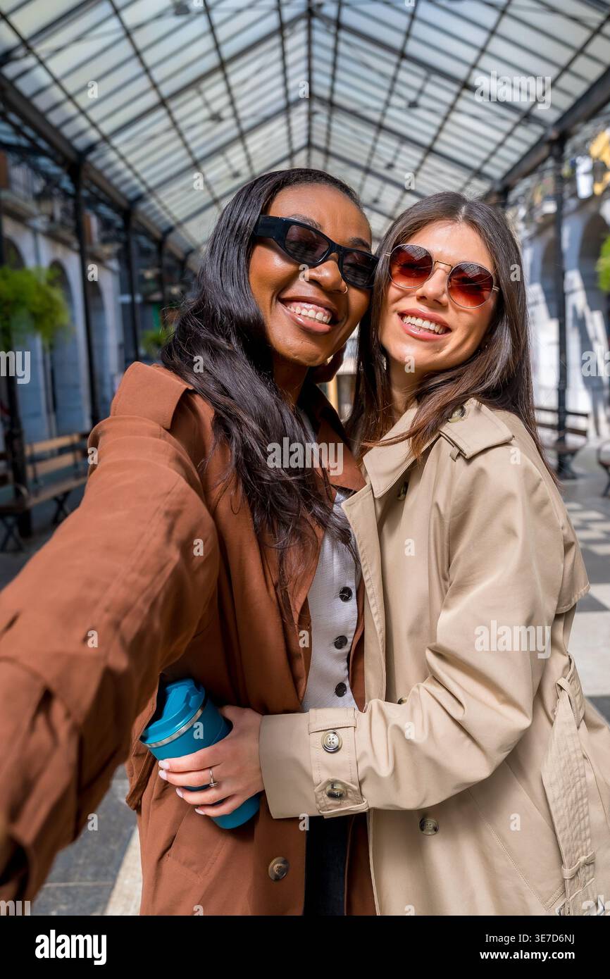 Amies noires et caucasiennes portant des trench-coat et des lunettes de soleil prenant des selfies tout en restant debout en ville pendant la journée Banque D'Images