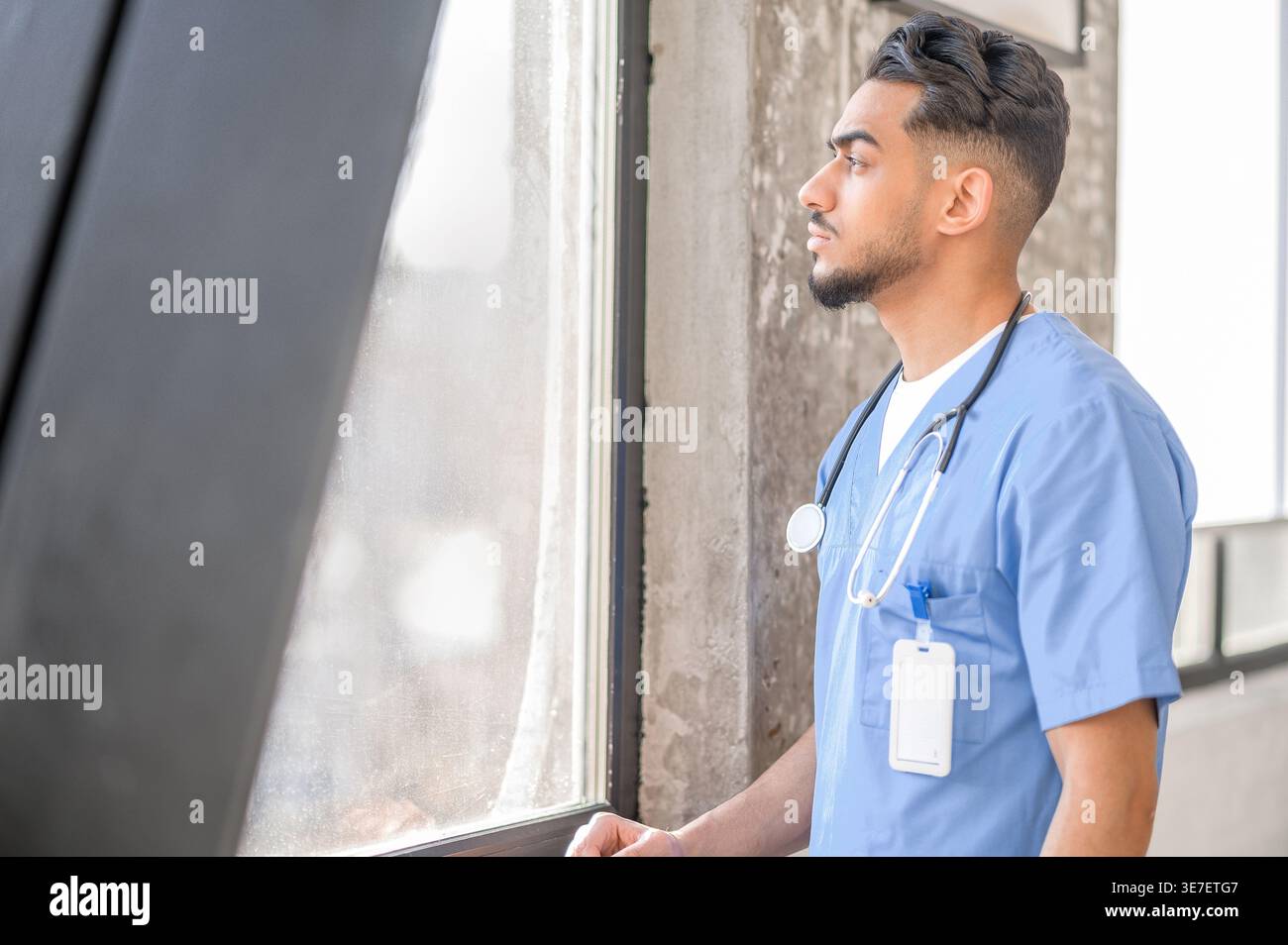 Médecin arabe sérieux avec stéthoscope sur le cou regardant loin debout près de la fenêtre à la maison Banque D'Images