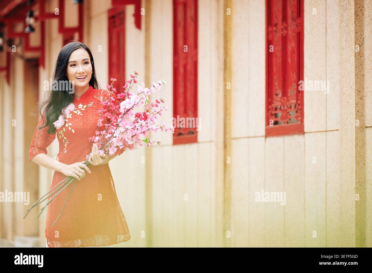 Femme asiatique souriante dans des vêtements traditionnels tenant des branches de pêche florissantes debout à l'extérieur Banque D'Images