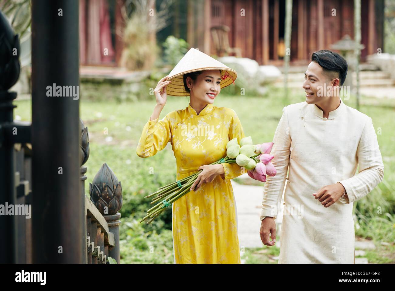 Femme asiatique dans des vêtements traditionnels portant un chapeau tenant des fleurs de lotus regardant son petit ami tout en marchant dans le jardin pendant la journée Banque D'Images