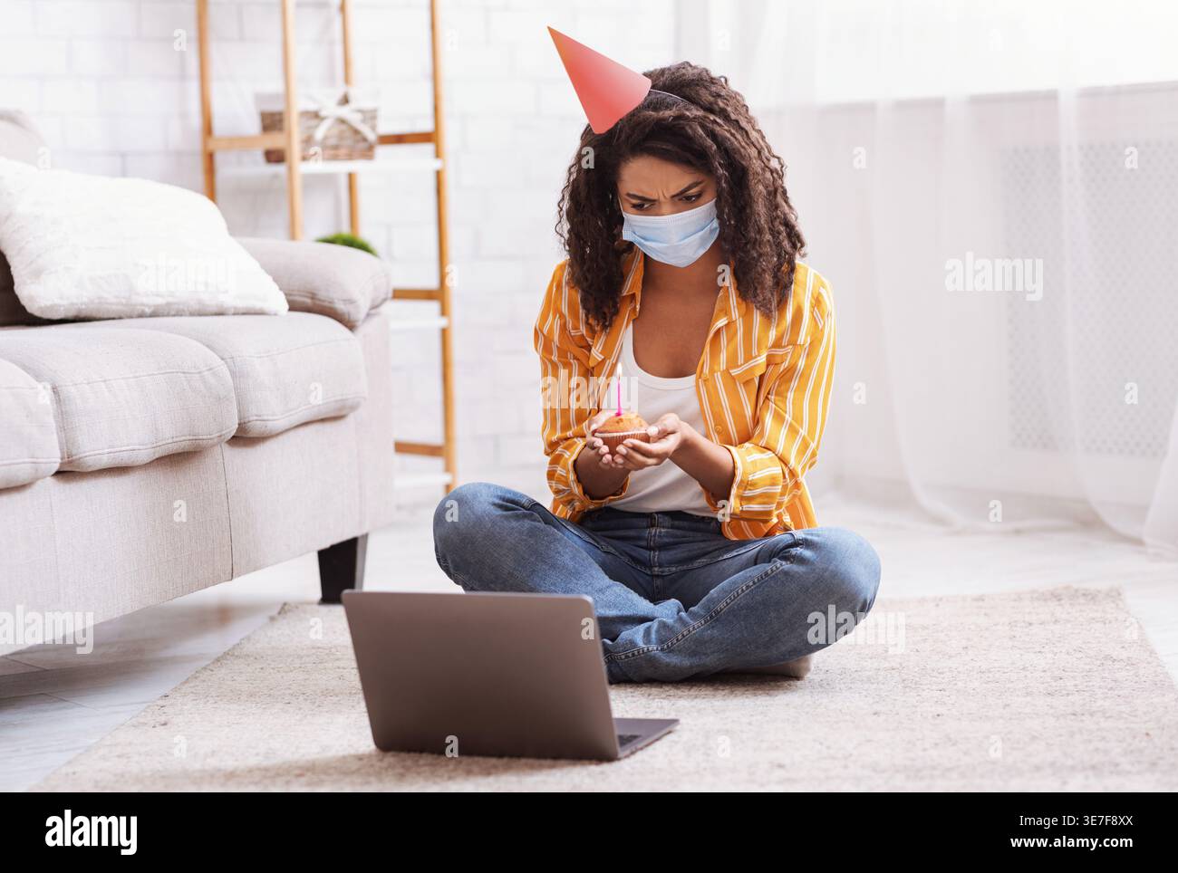 Contrarié femme noire portant chapeau de fête et masque médical tenant cupcake avec bougie et ayant appel vidéo sur ordinateur portable tout en étant assis sur la moquette du sol à la maison Banque D'Images