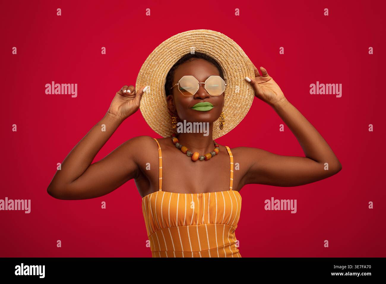 Femme noire avec les cheveux courts portant chapeau de paille et lunettes de soleil debout sur fond rouge dans le studio Banque D'Images