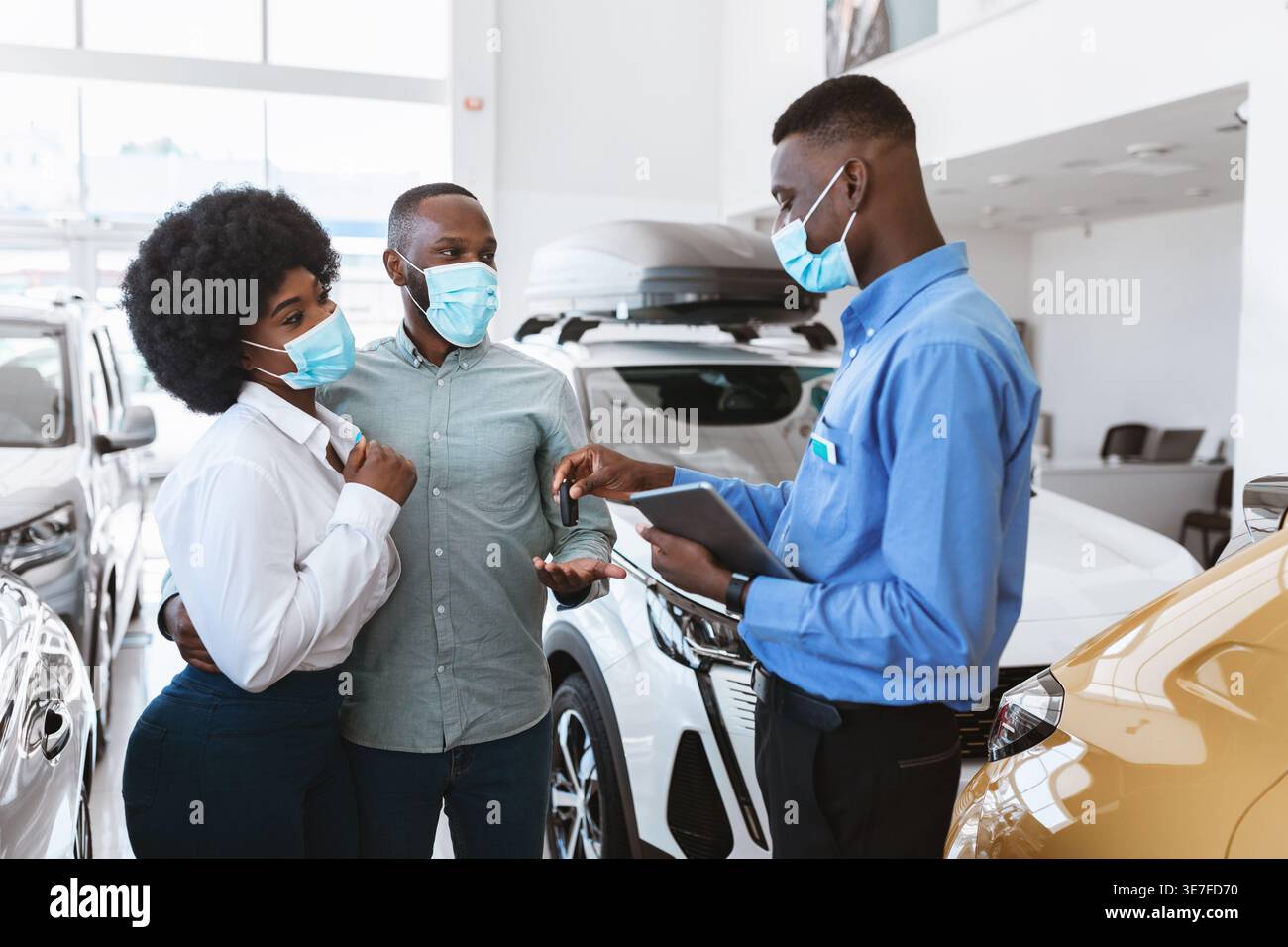 Couple noir dans les masques d'achat de nouvelle voiture et de prendre la clé du vendeur tenant la tablette tout en se tenant debout ensemble dans la salle d'exposition pendant la pandémie Banque D'Images