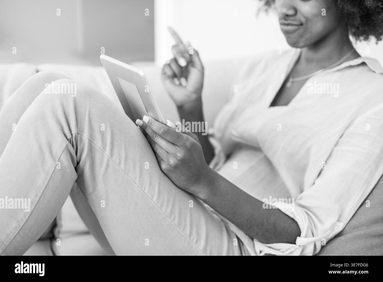 Image en noir et blanc de femme noire avec afro en utilisant la tablette assise avec les jambes sur le canapé dans le salon à la maison Banque D'Images
